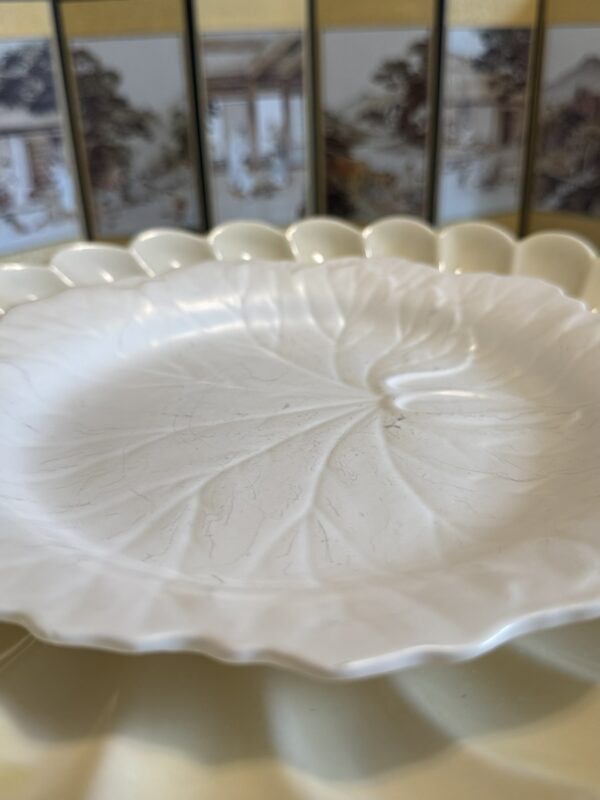 A white ceramic plate shaped like a lotus leaf sits on a pale yellow scalloped-edged dish. In the background, there is a decorative screen featuring traditional-style paintings of buildings and trees in muted colors.