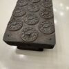 Rectangular stone block with twelve engraved clock faces showing different times. Each clock face features Roman numerals, and the block has a weathered, metallic appearance. The object sits on a pedestal against a plain, light-colored floor.