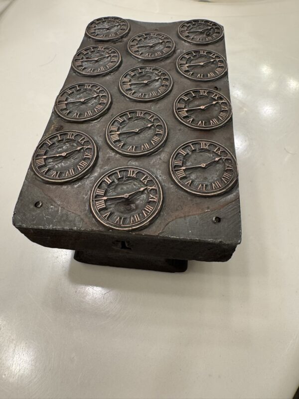 Rectangular stone block with twelve engraved clock faces showing different times. Each clock face features Roman numerals, and the block has a weathered, metallic appearance. The object sits on a pedestal against a plain, light-colored floor.