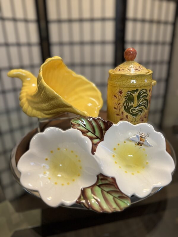 A ceramic dish shaped like two white flowers with green leaves and a painted bee rests on a tray. Behind it are a yellow cornucopia-shaped vase and a yellow canister with a red knob and green floral design. A folding screen is in the background.