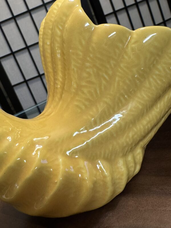 A close-up of a glossy yellow ceramic sculpture shaped like a stylized seashell, resting on a wooden surface. The background features a black grid room divider and beige panels. The ceramics surface shows embossed, textured patterns.