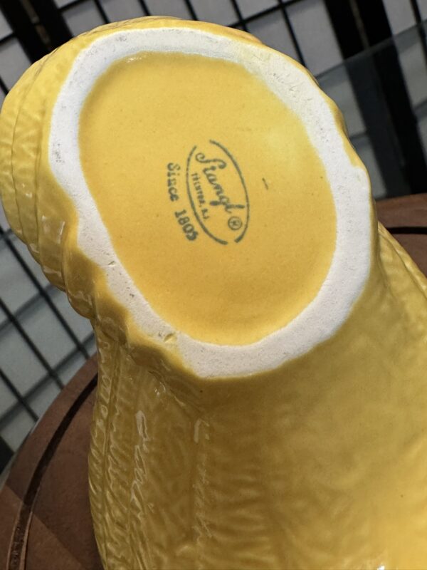 A close-up of the base of a yellow ceramic figure, showing the “François Since 1809” maker’s mark in black. The bottom rim is unglazed, exposing white ceramic. The background is blurred with vertical black bars and a brown surface.