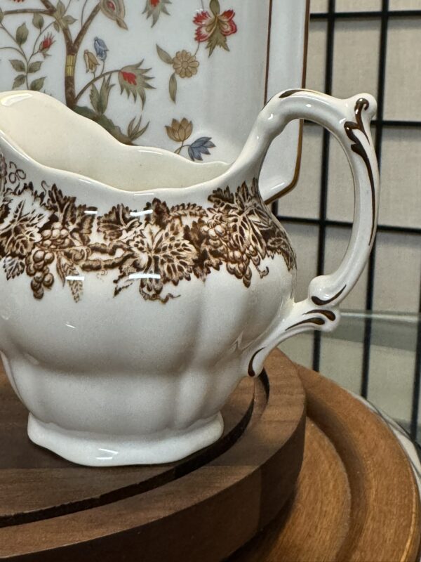 An ornate white ceramic creamer with a delicate brown floral pattern and a curved handle sits on a round wooden tray. Part of a rectangular plate with a colorful flower and branch design is visible in the background.