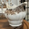 A white ceramic pitcher with a scalloped base and ornate brown floral and leaf patterns on its body and handle, sits on a wooden surface. Behind it, a white plate with a colorful floral design is partially visible.