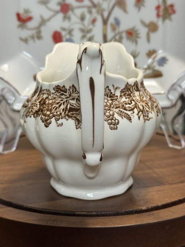 A vintage white ceramic creamer with a curved handle and brown floral and grapevine design sits on a wooden surface. In the background, floral-patterned dishes and a blurred floral wallpaper are partially visible.