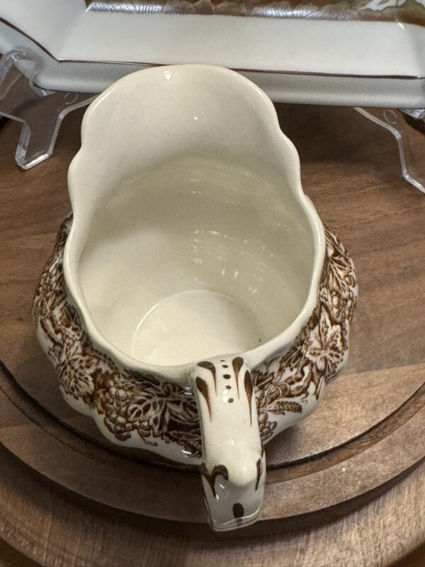 A vintage ceramic pitcher with a scalloped rim and ornate brown floral pattern sits on a wooden surface. The pitcher has a wide opening, a sturdy handle, and an off-white base color. A rectangular dish on a clear stand is partially visible in the background.