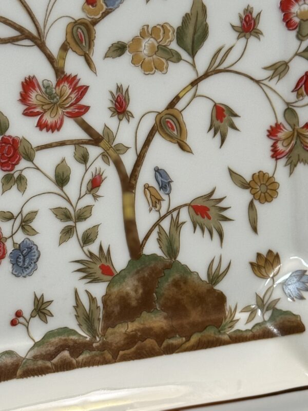 A close-up of a ceramic plate featuring a detailed floral design with red, yellow, and blue flowers, green leaves, and two small birds perched on a tree branch above a rocky mound, all outlined in gold. The background is white.