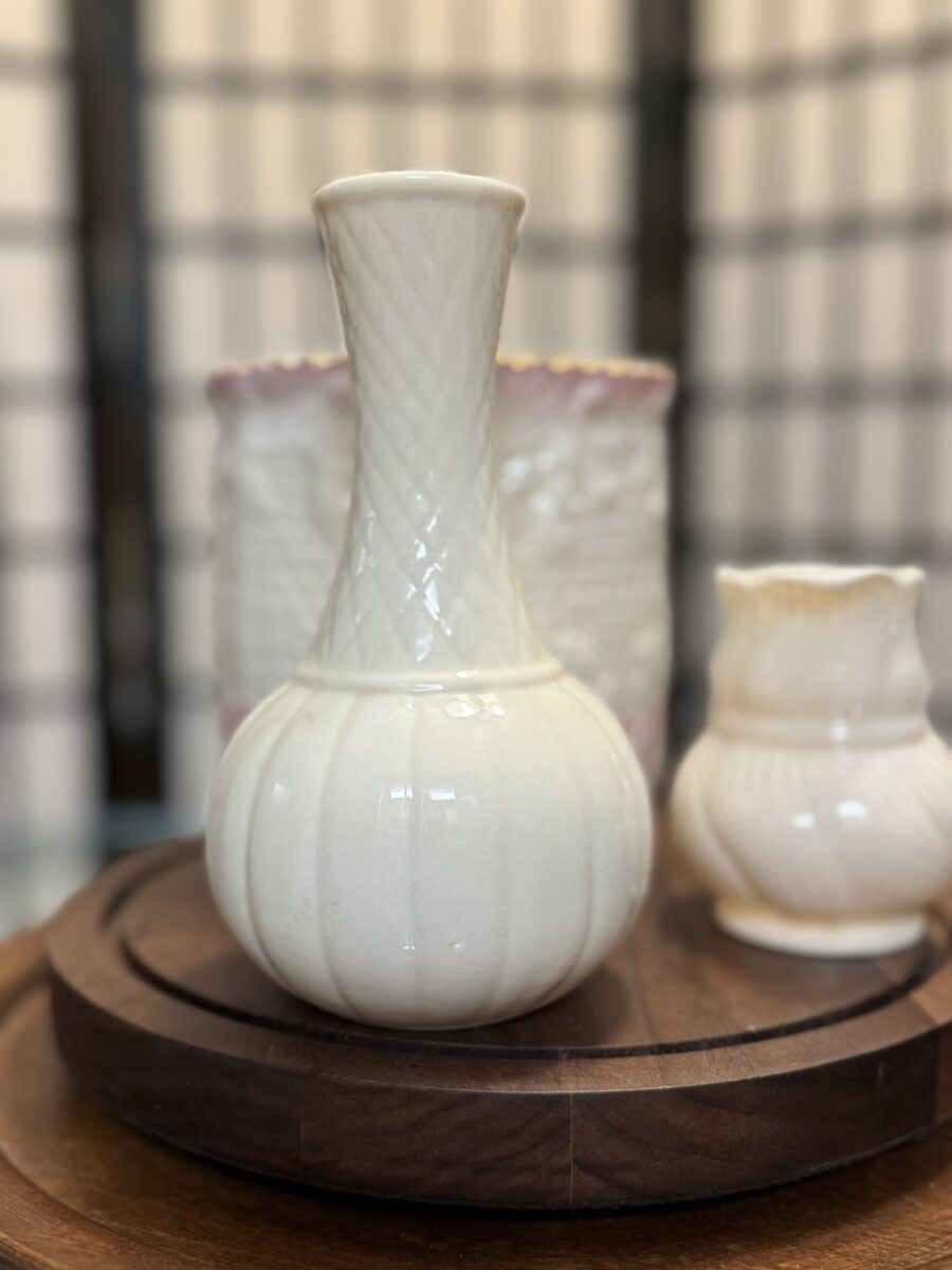 A white ceramic vase with a rounded base and narrow, textured neck sits on a wooden tray. Two other small, cream-colored vases are partly visible in the background. The scene is softly lit, with a blurred, grid-patterned backdrop.