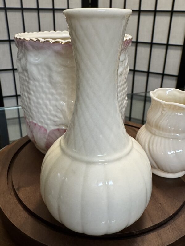 A glossy, cream-colored ceramic vase with a bulbous base and narrow neck sits on a wooden tray. Behind it are two other ceramic pieces: a scalloped-edge pot with raised texture and pink accents, and a smaller, ribbed cream-colored vase.