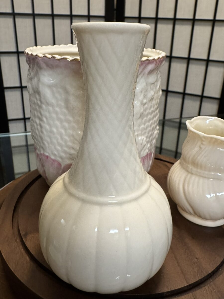 A cream-colored ceramic vase with a bulbous base and narrow neck sits on a wooden surface. Behind it are a textured, scalloped-edge bowl with pink accents and a small, ribbed cream pitcher. A black grid screen forms the background.