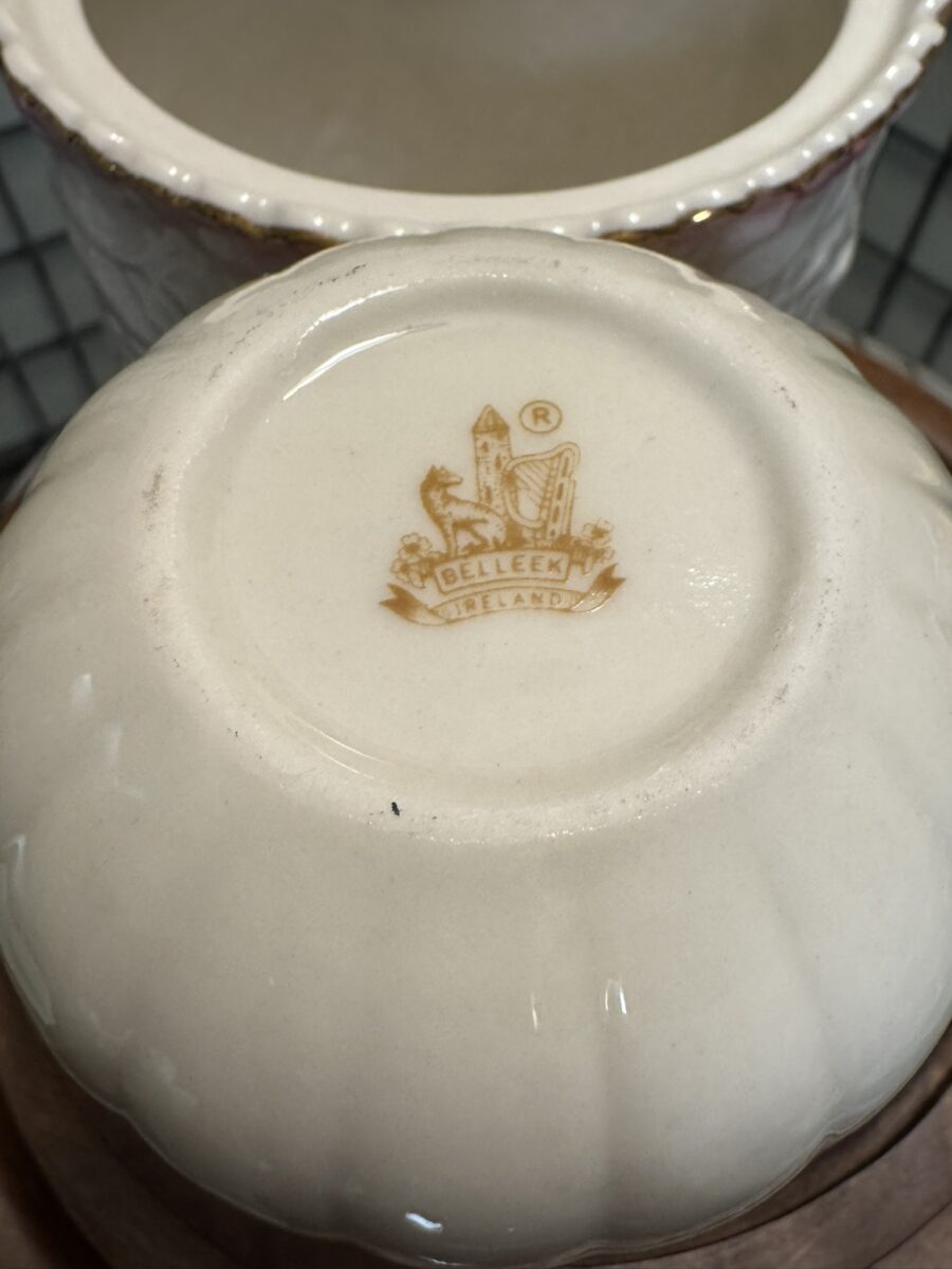 A close-up of the bottom of a cream-colored ceramic bowl featuring the Belleek Ireland logo in brown, with a harp, castle, and wolfhound design above the text BELLEEK IRELAND. The bowl has subtle ridges and a glossy finish.