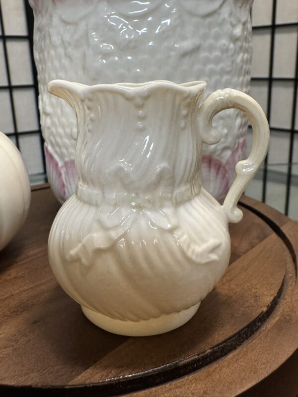 A cream-colored, ornate ceramic pitcher with a wavy rim, decorative handle, and embossed vine-like details sits on a wooden tray. The background shows a textured, white and pink ceramic vase and a black grid. The pitcher has a glossy finish.