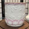 A white ceramic cup with a scalloped gold rim, textured surface resembling fish scales, and embossed seashells around the base tinted with pink. The cup sits on a wooden surface, with a blurred, black-and-white paneled screen in the background.