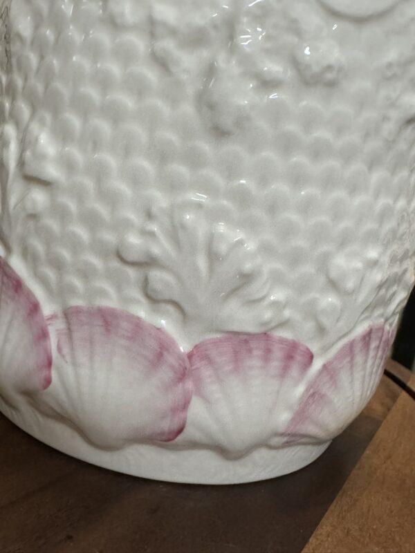 A close-up of a white ceramic vase featuring embossed textured patterns and a row of scallop shell designs near the base, accented with pale pink edges. The vase sits on a wooden surface.