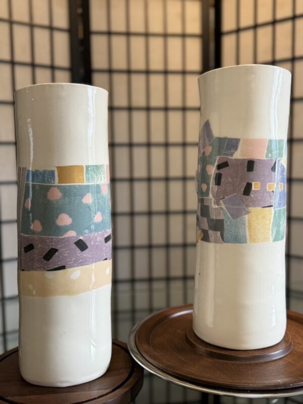 Two tall, glossy white ceramic vases stand side by side on wooden trays. Each vase features a colorful geometric pattern with pastel shapes in blue, pink, yellow, purple, and black, set against a blurred grid-patterned room divider background.