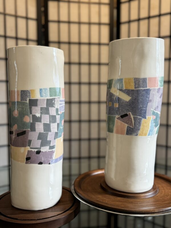 Two tall, white ceramic vases sit on wooden stands. Each vase features a colorful, abstract geometric pattern with pastel hues of green, pink, yellow, and blue. A folding screen with a grid pattern is blurred in the background.