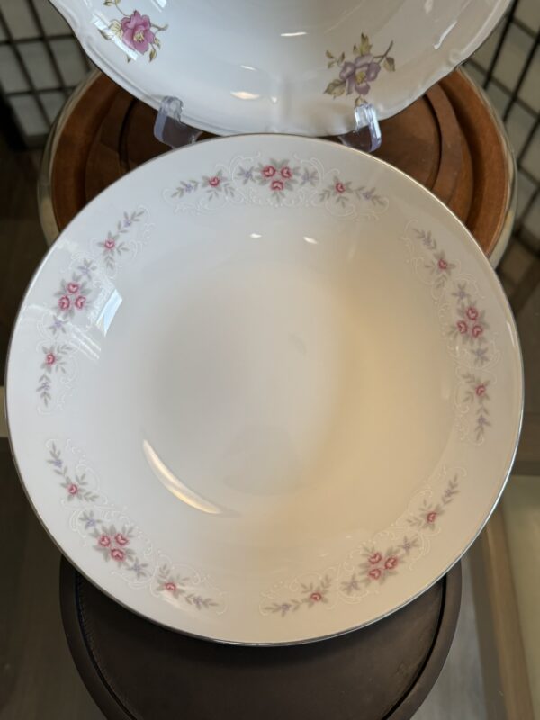A white porcelain bowl with a subtle embossed floral pattern and decorated with small pink and lavender flowers along the rim. The bowl is displayed on a round wooden surface, with part of another matching dish visible in the background.