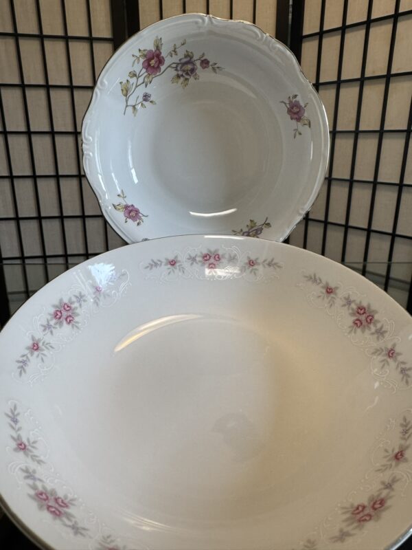 Two white porcelain dishes with floral designs are displayed. The front dish is large with light pink and white flowers along the rim. The back dish is smaller, featuring delicate pink and yellow flowers with green leaves around the edge. A black grid is behind them.