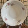A cream-colored ceramic bowl with scalloped edges and pink and yellow floral designs sits on a clear plastic stand. The background features a wooden folding screen with a grid pattern. The bowl rests on a round wooden surface.