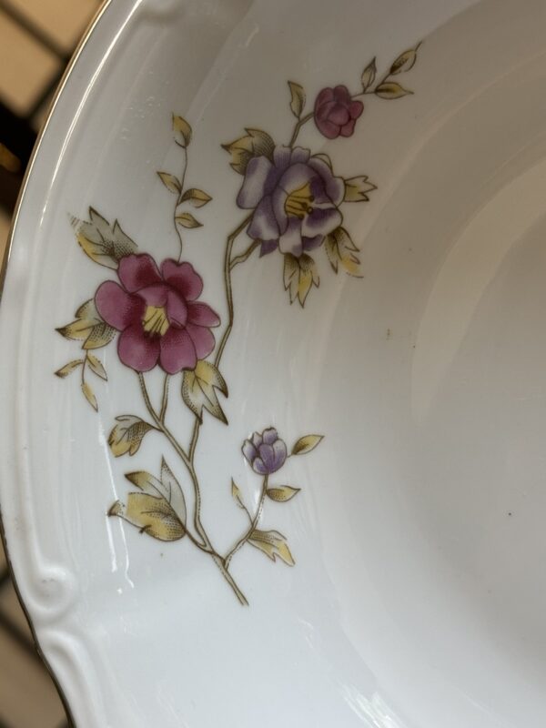 A close-up of a white porcelain plate with a floral design featuring pink and purple flowers with yellow centers and greenish-yellow leaves on delicate stems, set against the plate’s glossy surface with a scalloped rim.
