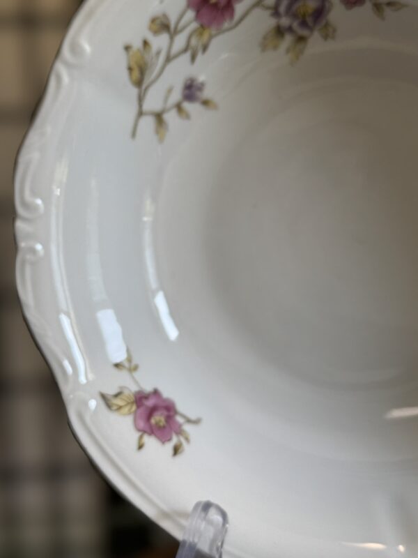 A close-up of a white porcelain plate with embossed edging, decorated with pink and yellow floral patterns along the rim. The plate sits on a clear stand, and the background is softly blurred.
