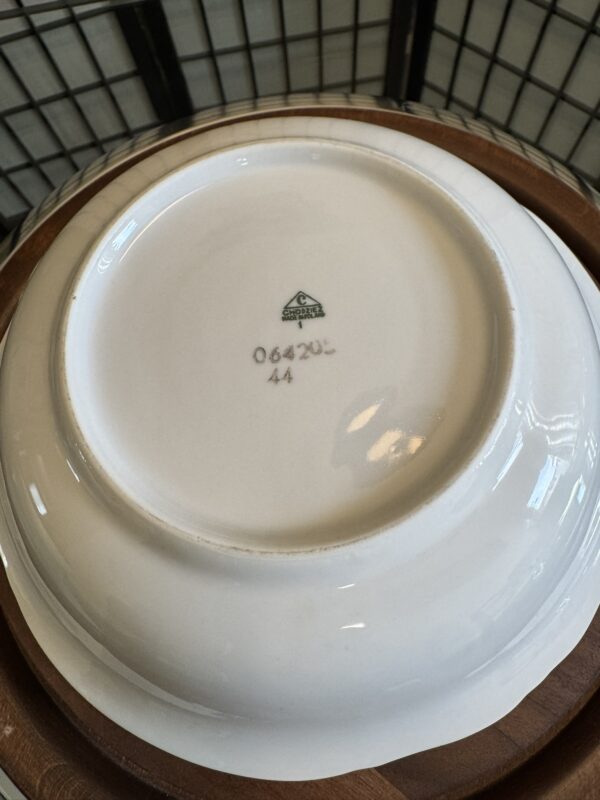 A white ceramic bowl upside down, showing its underside. At the center is a green stamp with a triangular logo and the word “Syracuse” underneath, along with the numbers “064 201” and “44.” The bowl sits on a wooden tray with a wire grid background.