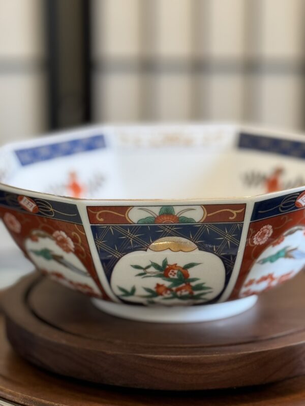 An ornate porcelain bowl with octagonal edges, featuring intricate red, blue, and gold designs of flowers and traditional motifs on its outer surface, sits on a round wooden base against a softly blurred background.