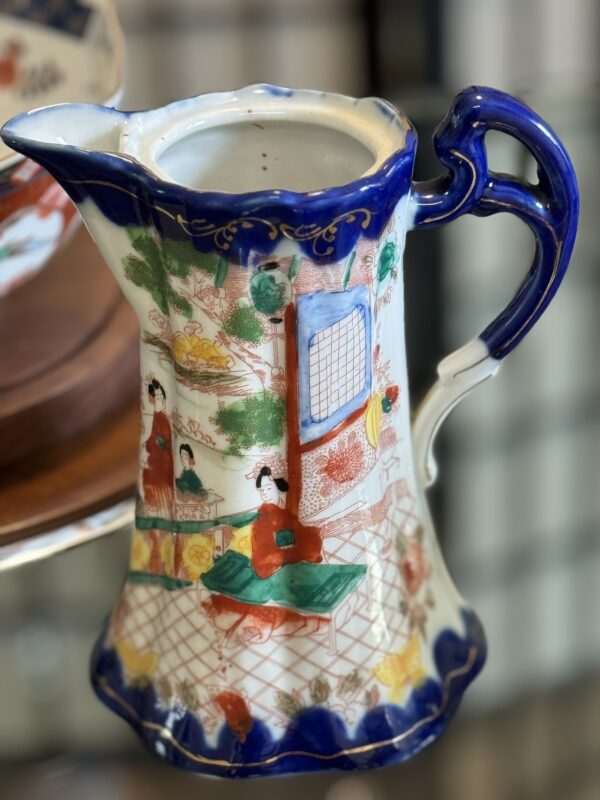 A porcelain pitcher with a fluted rim and blue handle, decorated with colorful, hand-painted Asian-inspired scenes featuring people in traditional attire, a lattice pattern, and accents of gold, blue, red, and green. The background appears softly out of focus.