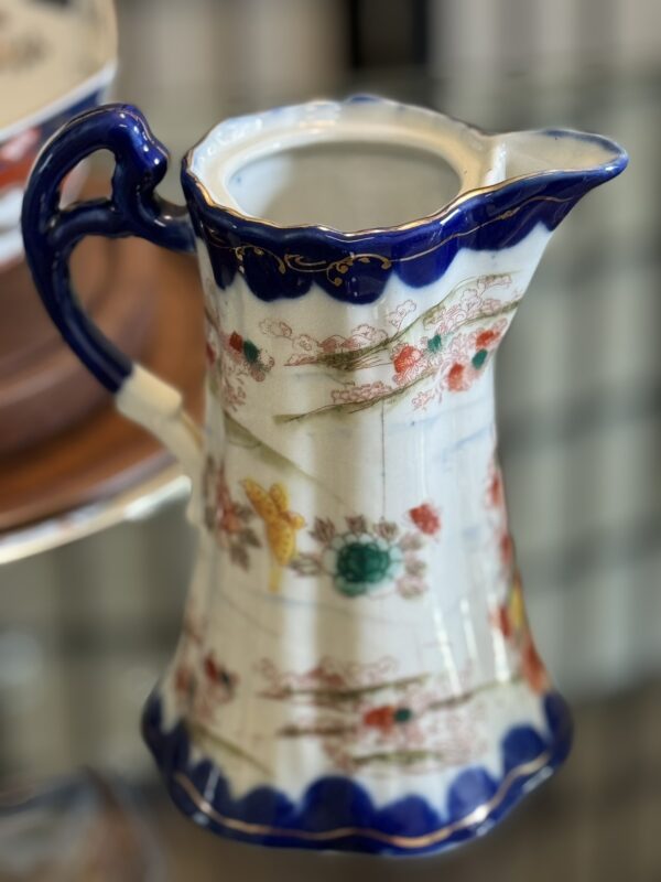 A ceramic pitcher with a blue scalloped rim and handle, decorated with intricate floral patterns in red, green, and gold. The body is cream-colored, adorned with delicate, painted flowers and gold accents, sitting on a glass shelf with a blurred background.