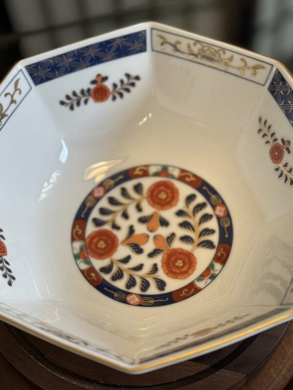 A hexagonal porcelain bowl with intricate floral and geometric patterns in blue, orange, and gold. The bowls center features three orange flowers with blue leaves, surrounded by decorative borders and leaf motifs along the inner sides.