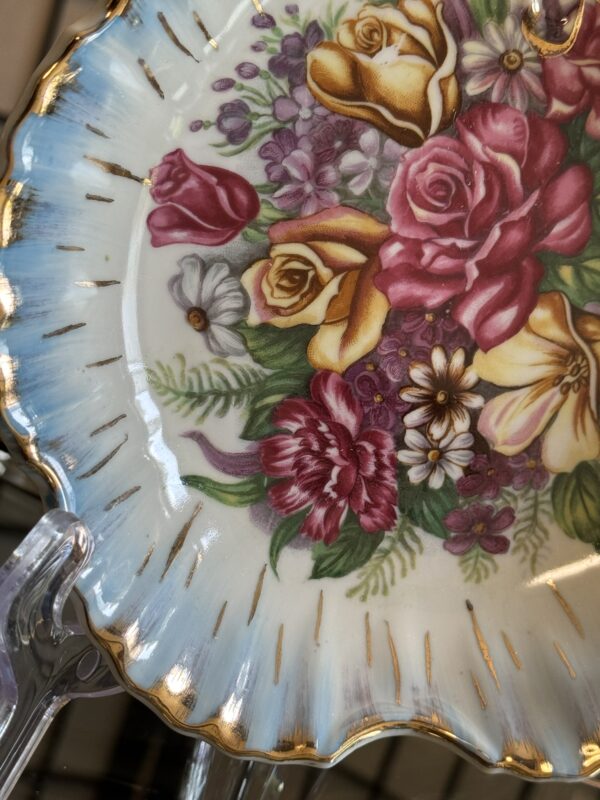 A decorative porcelain plate with a scalloped edge, blue and gold trim, and a detailed floral design featuring pink, yellow, and red roses, purple flowers, and green leaves. The plate rests on a clear display stand.