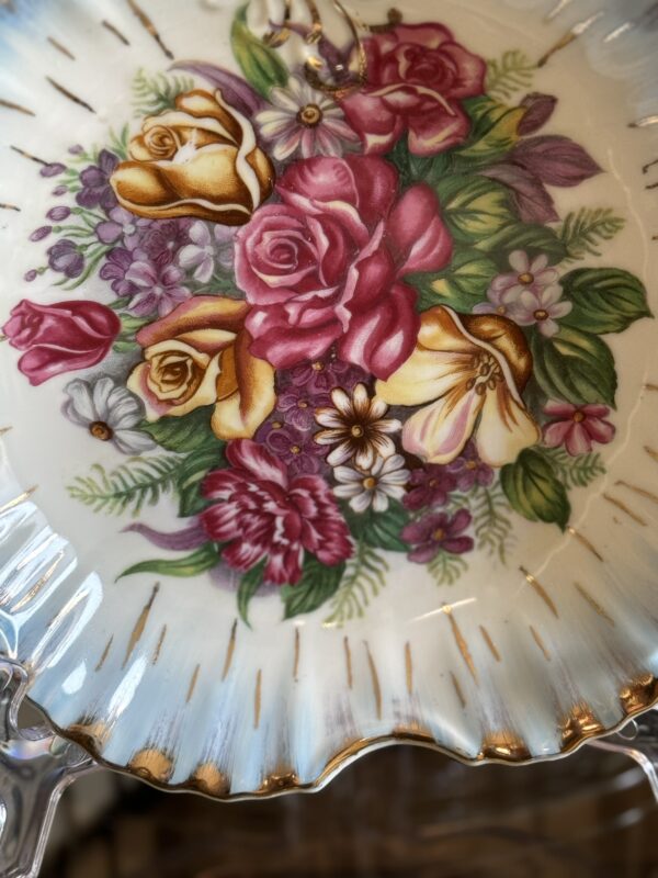 A decorative ceramic plate with scalloped, gold-trimmed edges features a vibrant floral design. The bouquet includes pink roses, yellow flowers, purple and white blossoms, and green leaves, set against a cream background with golden streak accents.