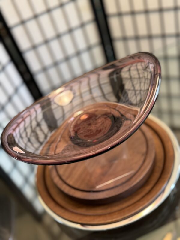 A translucent, purple-tinted glass bowl with a wide rim sits on a wooden base. The background features a blurred folding screen with a grid pattern in neutral tones. Warm light reflects off the bowl’s smooth surface.