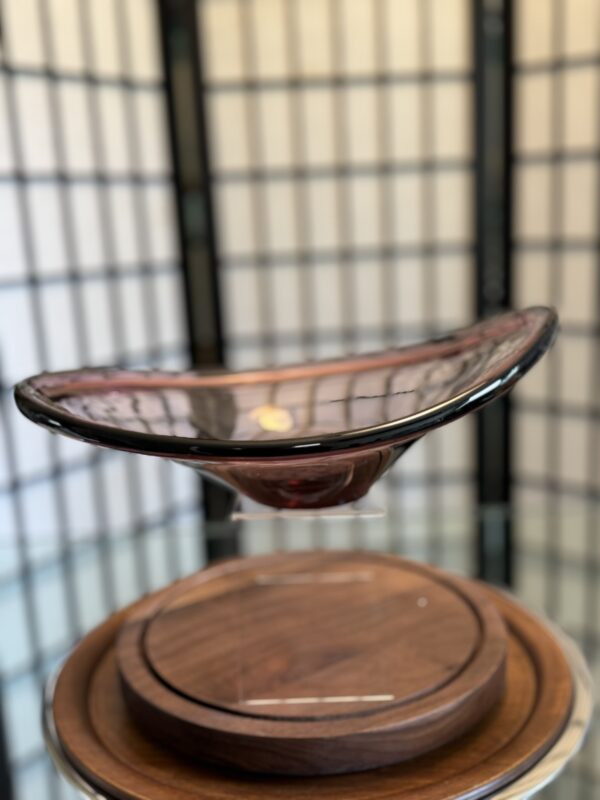 A shallow, translucent glass bowl with a pinkish hue, featuring a wavy rim and irregular shape, sits elevated on a clear stand above a round wooden base. The background consists of a softly focused grid-patterned privacy screen.