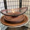 A decorative glass bowl with a curved, translucent reddish rim sits atop stacked round wooden trays and a plate, displayed on a glass shelf. A folding screen with a grid pattern is blurred in the background.