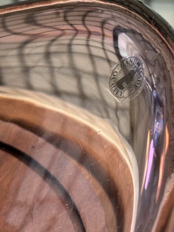 Close-up of a transparent, slightly purple glass object with curved edges. A round sticker saying “Handmade” is visible on the glass. The background shows a reflected grid pattern and warm-toned, blurry shapes.