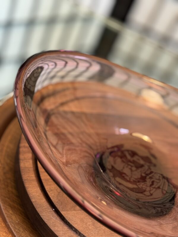 A close-up of a transparent, pink-tinted glass bowl on a circular wooden tray. The focus is on the bowl’s curved edge, with reflections and light patterns visible. In the background, there are blurred lines from a window or railing.