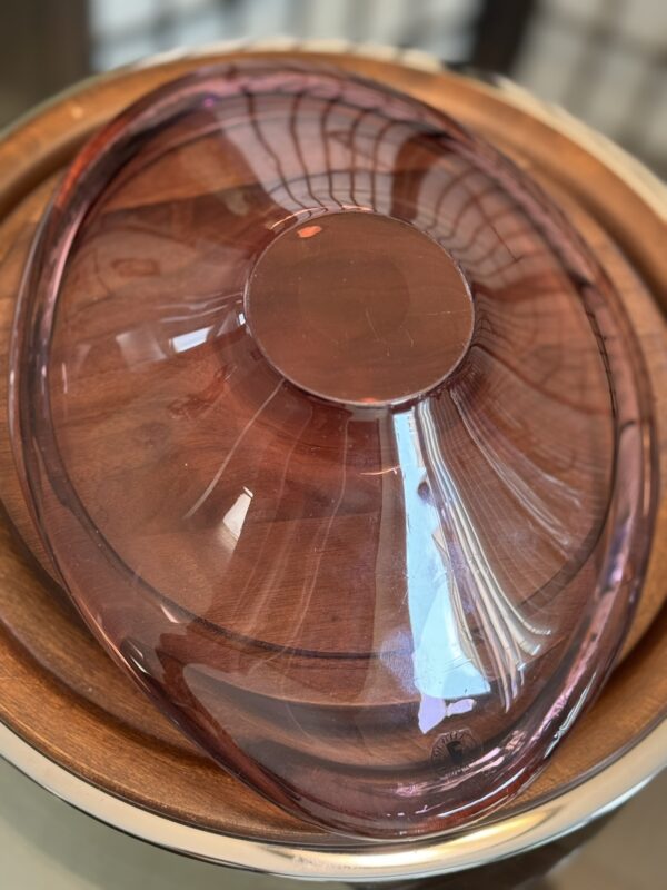 A translucent, oval-shaped pink glass bowl is placed upside down on a smooth wooden tray. Reflections of a window grid and surrounding light are visible on the shiny surface of the bowl. The background is softly blurred.