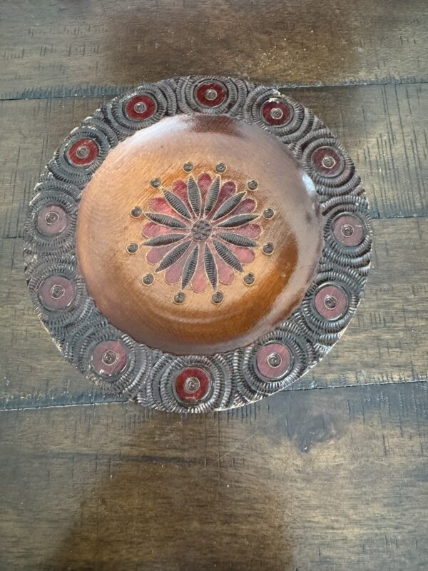 A round, carved wooden plate with a floral design in the center and a decorative border of red circles and textured carvings, sits on a dark, rustic wooden table. The plate features a polished finish and intricate detailing.