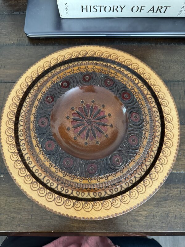 A wooden plate with intricate hand-carved patterns in concentric circles, featuring dark brown, red, and gold designs. The center has a flower-like motif. The plate sits on a wooden table, with a History of Art book partially visible above it.