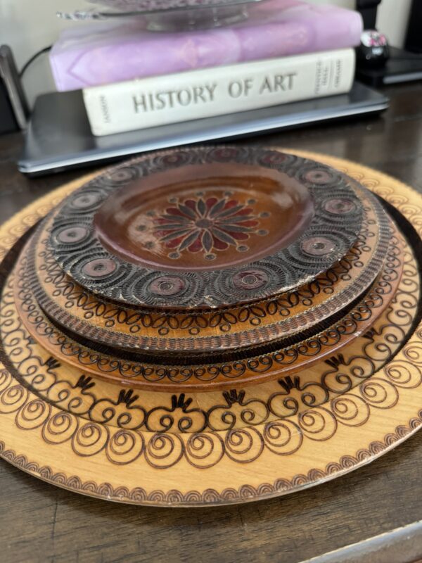 Four intricately patterned plates are stacked on a dark wood surface. Each plate features different brown and black ornamental designs. In the background, there is a white HISTORY OF ART book and a lavender item resting on top of it.
