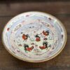 A decorative ceramic plate sits on a wooden surface. The plate features colorful hand-painted roosters, green grass, and red flowers in the center, surrounded by intricate blue, red, and gold patterns along the rim.