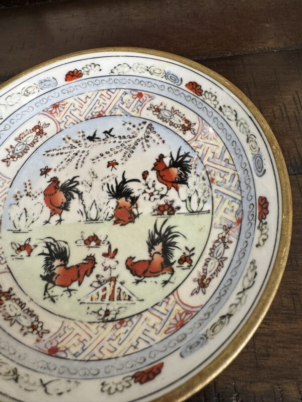 A decorative porcelain plate with a gold rim features a central scene of four red and black roosters among flowers and grass, surrounded by intricate red, blue, and gold patterns and floral motifs on a cream background. The plate rests on a wooden surface.
