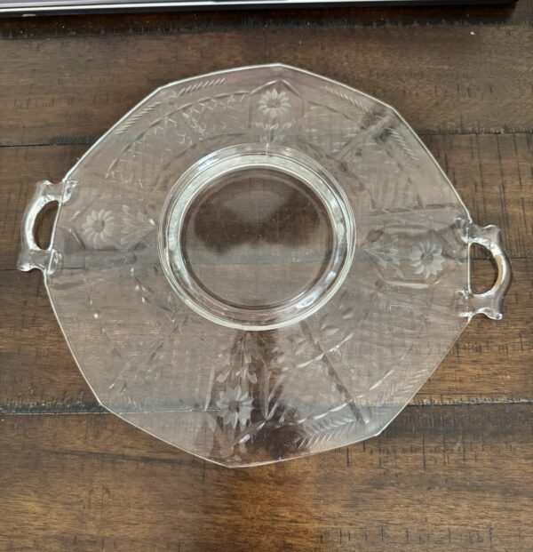 A clear, octagonal glass serving tray with two small handles and etched floral patterns sits on a wooden table next to a book titled History of Art. The trays surface reflects light, highlighting its intricate design.