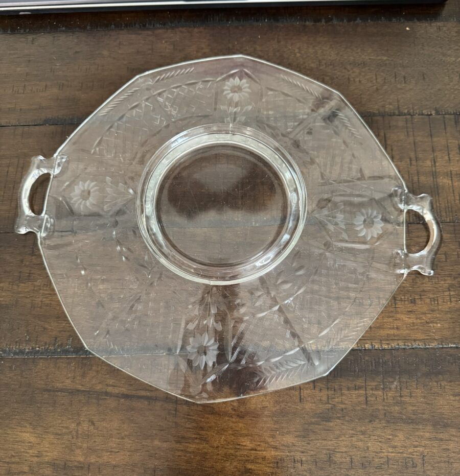 A clear, octagonal glass serving tray with two small handles and etched floral patterns sits on a wooden table next to a book titled History of Art. The trays surface reflects light, highlighting its intricate design.