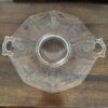 A clear, octagonal glass serving dish with two ornate handles and etched leaf patterns sits on a wooden table. The dish has a circular center and geometric designs. A closed laptop and a book titled History of Art are visible in the background.