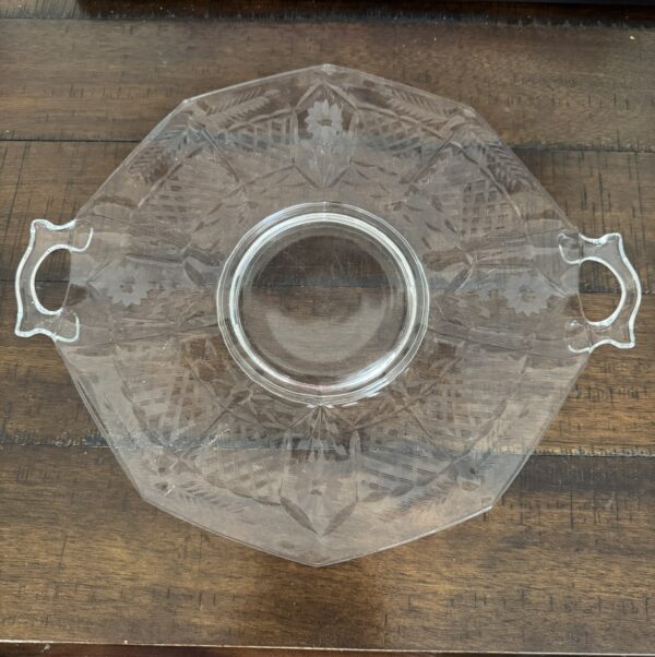 A clear, octagonal glass serving dish with two ornate handles and etched leaf patterns sits on a wooden table. The dish has a circular center and geometric designs. A closed laptop and a book titled History of Art are visible in the background.