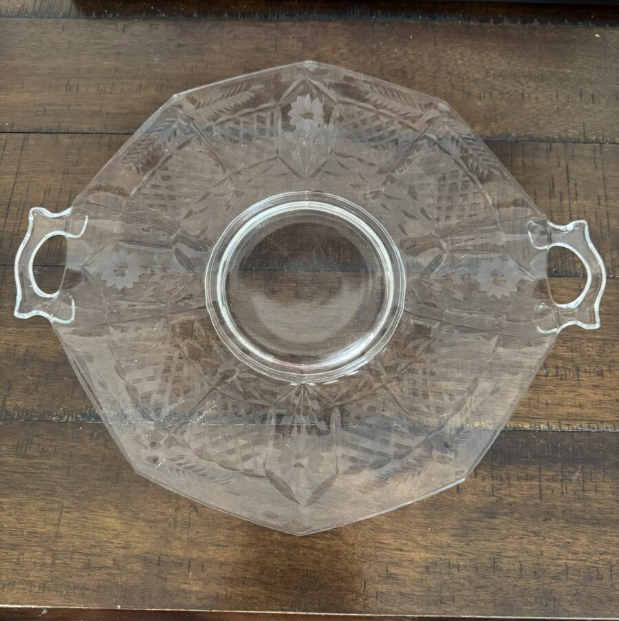 A clear, octagonal glass serving dish with two ornate handles and etched leaf patterns sits on a wooden table. The dish has a circular center and geometric designs. A closed laptop and a book titled History of Art are visible in the background.