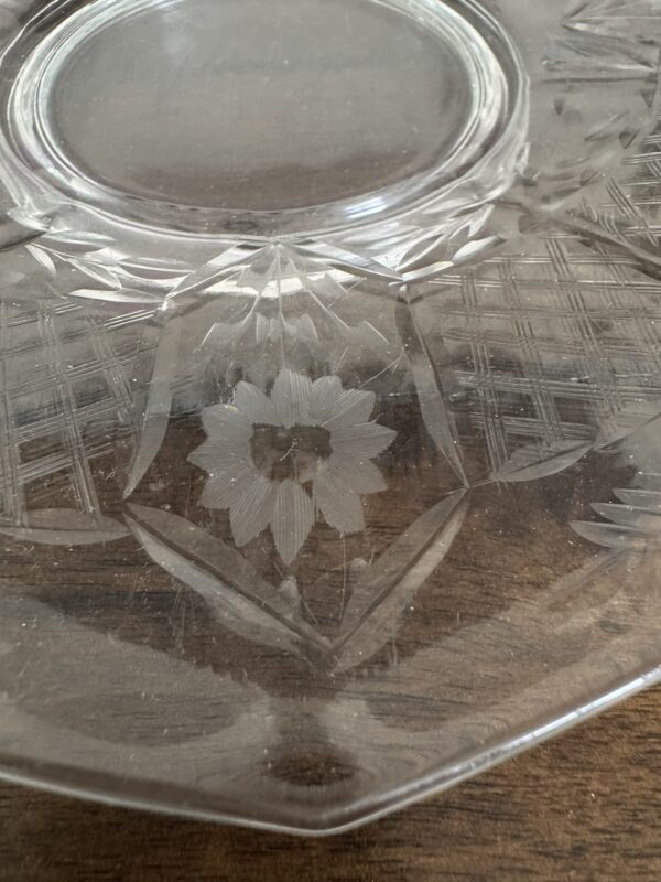 A close-up of a clear glass plate with an etched floral design in the center and decorative geometric patterns around it. The plate rests on a brown wooden surface. The details of the etching are visible and intricate.
