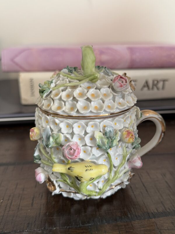 The Meissen Schneeballen Cup is a small, ornate porcelain cup with a handle, featuring raised white flowers, pink roses, a yellow bird, and a lid topped with floral details. It rests on wood with blurred books in the background.